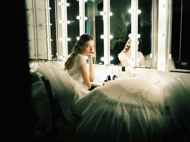 Young woman in a white dress sits in front of a lighted vanity mirror, surrounded by tulle fabric in a dressing room setting.