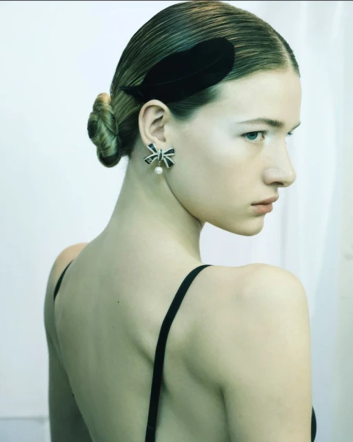 Elegant portrait of a young woman with bare shoulders, wearing a black bow earring and a black feather hair clip, against a white backdrop.