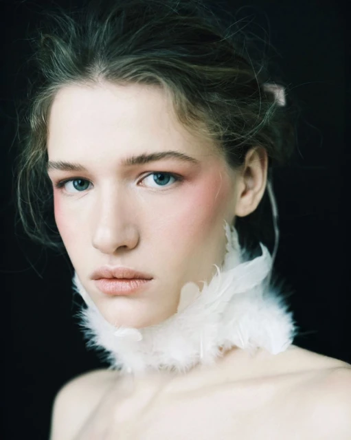 Close-up portrait of a young woman with fair skin and blue eyes, accented with pink blush, posing with a white feathered collar against black.