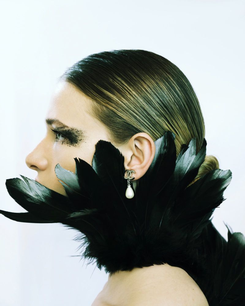 Profile view of a person with dramatic black glitter eyeshadow and a black feather collar with a dangling pearl earring on white.