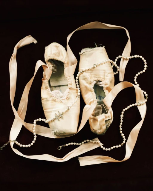 Worn ballet pointe shoes with pearl necklace and pale pink ribbons on dark surface for dance, fashion, or theater themes.