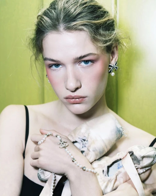 Close-up shot of a fair-skinned adult woman with light blonde hair, blush, pearl jewelry, and black top, against a light green background.