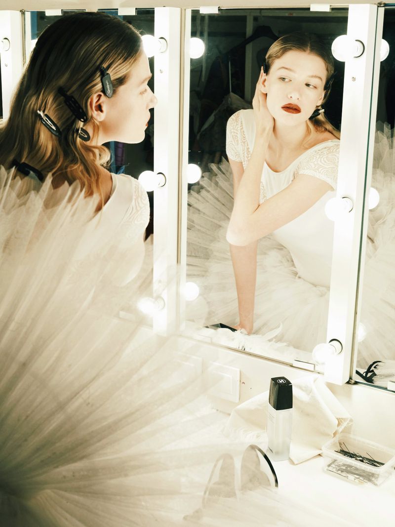 Elegant woman with fair skin in a white dress looks in a vanity mirror with lights in an indoor setting, applying makeup.