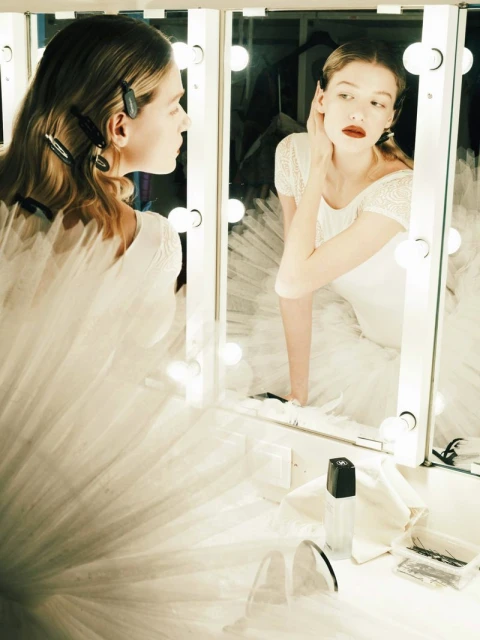Elegant woman with fair skin in a white dress looks in a vanity mirror with lights in an indoor setting, applying makeup.