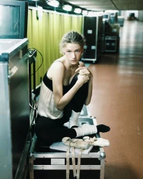 Model Mila Prvulovic seated on an equipment flight case backstage wearing a camisole top, tights, leg warmers, and ballet shoes.