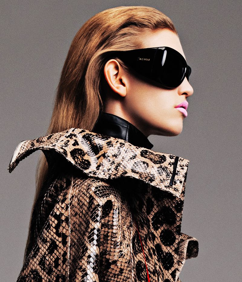 A stylish woman with blonde hair poses in sunglasses and a snakeskin-print jacket against a grey background for a fashion or beauty editorial.