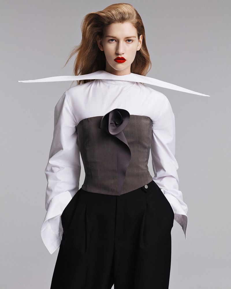 A fair-skinned woman with red lipstick poses in a white blouse with a gray corset and black pants in a fashion studio setting.