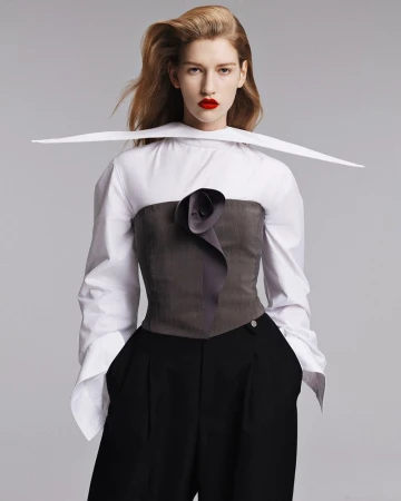 A fair-skinned woman with red lipstick poses in a white blouse with a gray corset and black pants in a fashion studio setting.