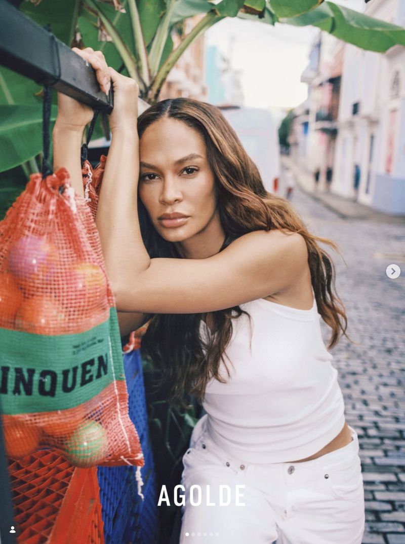 Adult woman with long brown hair poses in front of a blurred city street, holding onto a metal bar with fruit in a mesh bag.