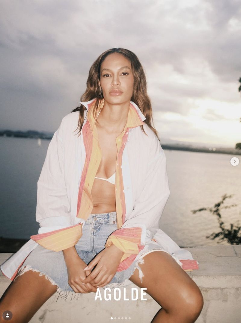 A woman poses in an unbuttoned white shirt and jean shorts against a cloudy outdoor seascape at dusk, showcasing fashion and style.