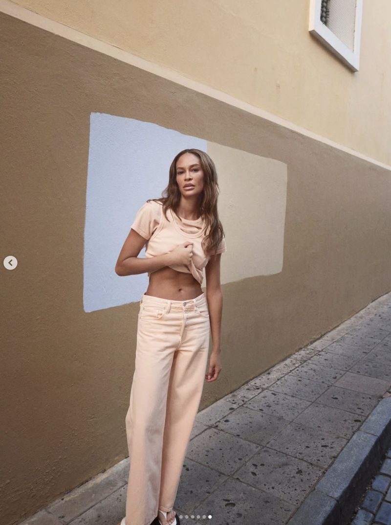 Model Joan Smalls poses on an urban sidewalk wearing a light peach t-shirt and matching wide-leg trousers against a painted wall.