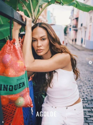 Model Joan Smalls leaning against a rail outdoors with a mesh bag of oranges in AGOLDE campaign