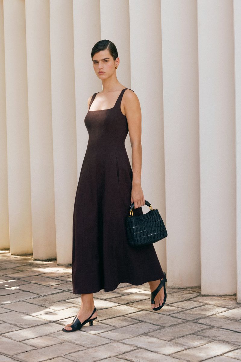 Brunette woman in a brown sundress, strappy sandals, and black purse walking in an outdoor setting with concrete columns.