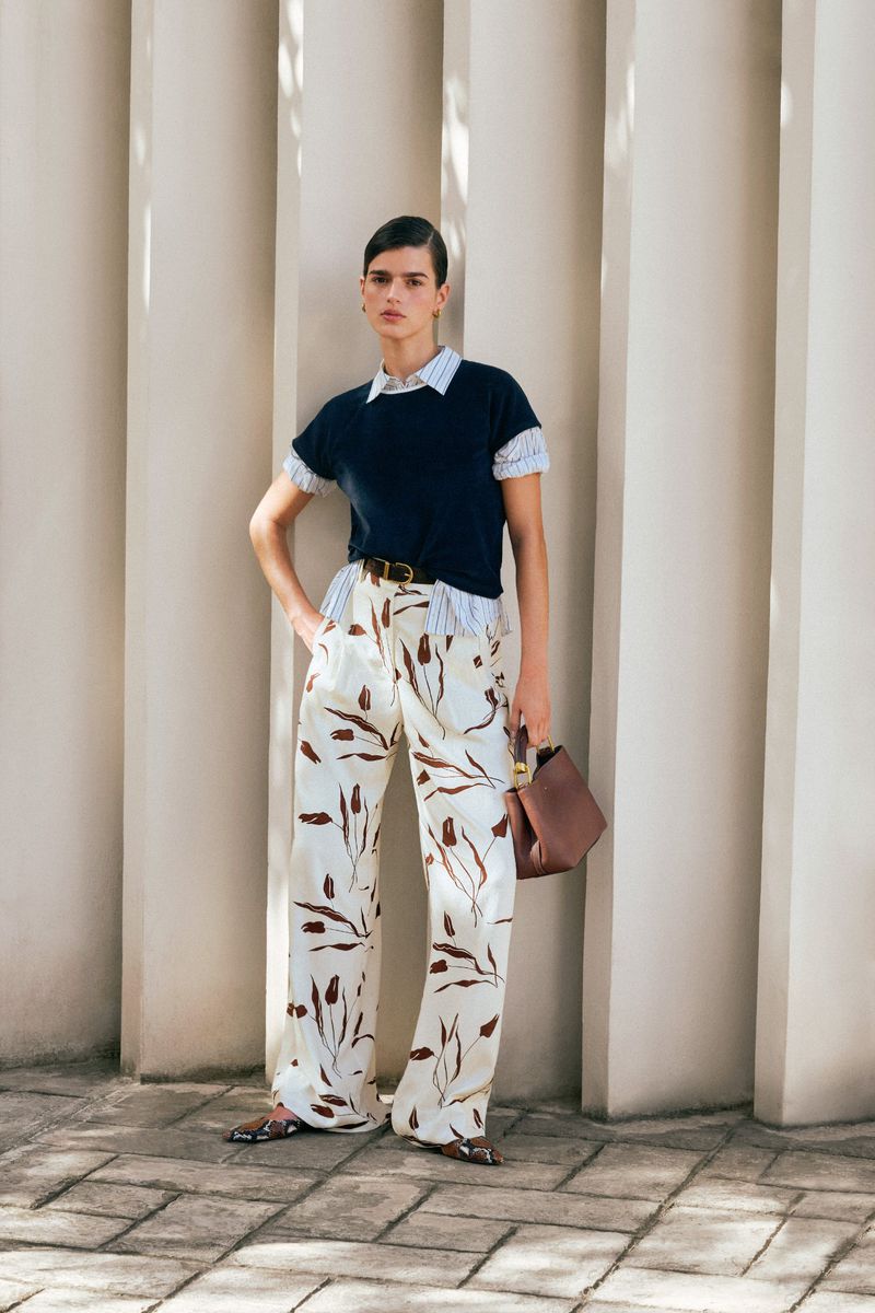 A woman stands in casual fashion with white patterned pants and navy shirt, against a textured neutral background with vertical elements.