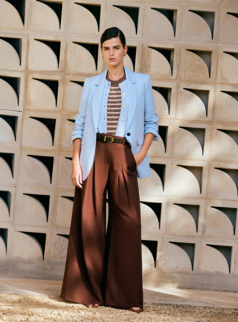 Fashion shot of a woman in a blue blazer and brown pants, standing against a geometric block wall with light and shadow.