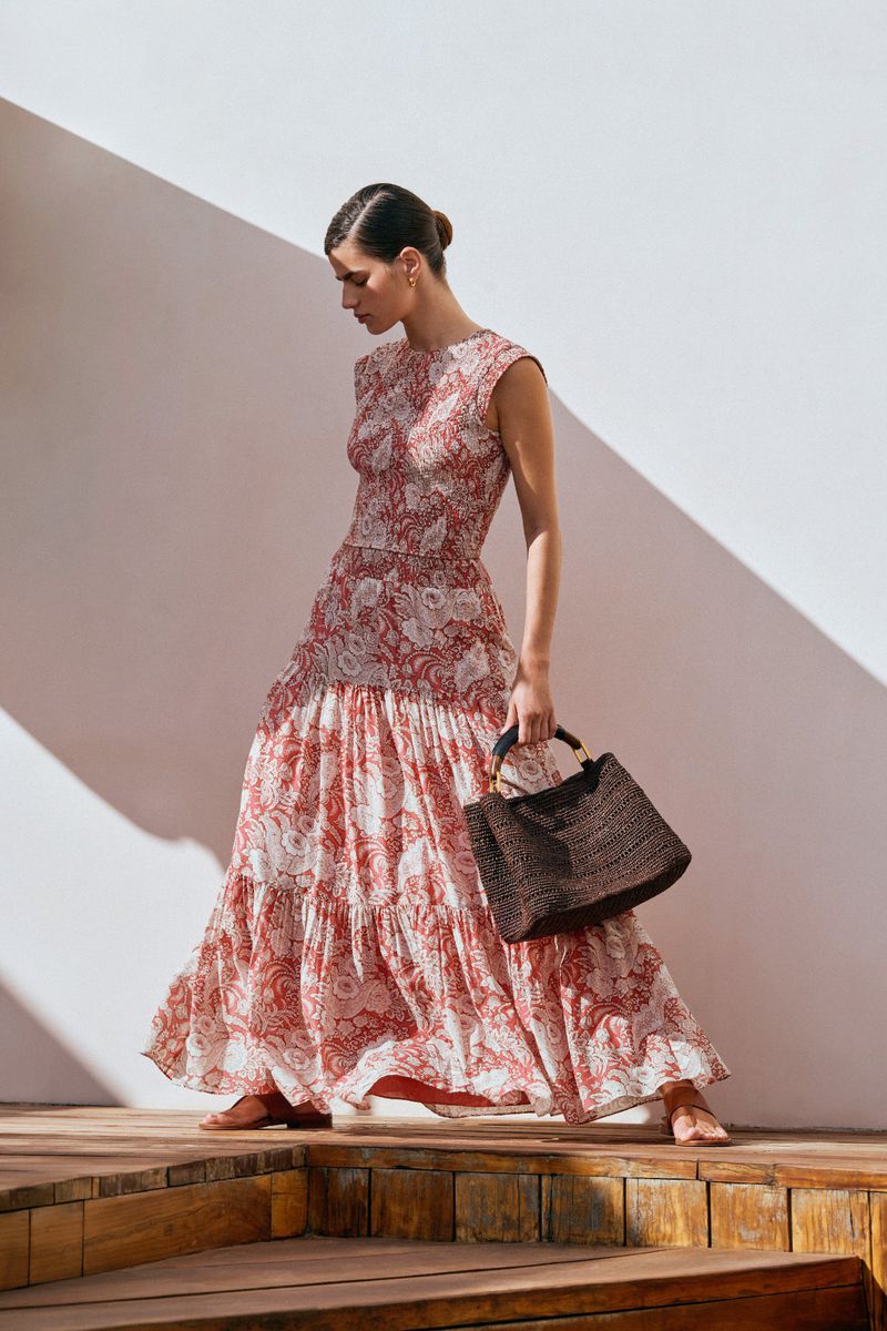 Woman in patterned maxi dress with a woven handbag, standing on wood steps against a white wall and a sunlight shadow.