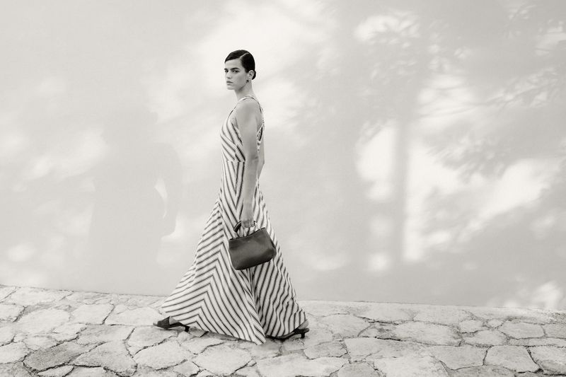 A female model walks on a stone path, carrying a purse, with a sunlit wall and tree shadows behind her in a fashion editorial image.