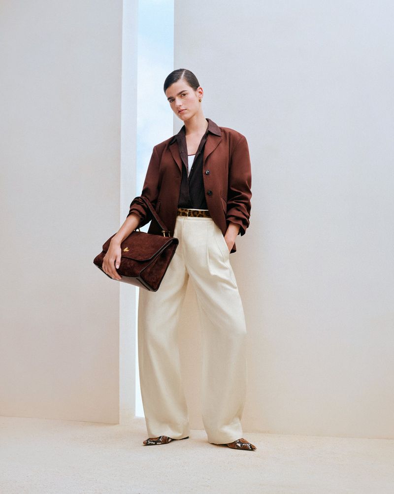 A woman poses indoors wearing a brown blazer, white trousers, and carrying a brown purse, bathed in bright lighting.