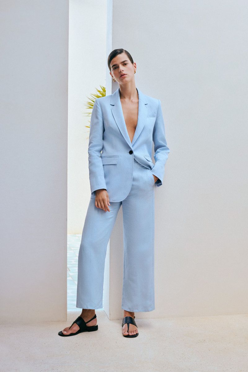 Woman in a light blue suit and black sandals stands indoors with a neutral backdrop, exuding a relaxed and stylish vibe.