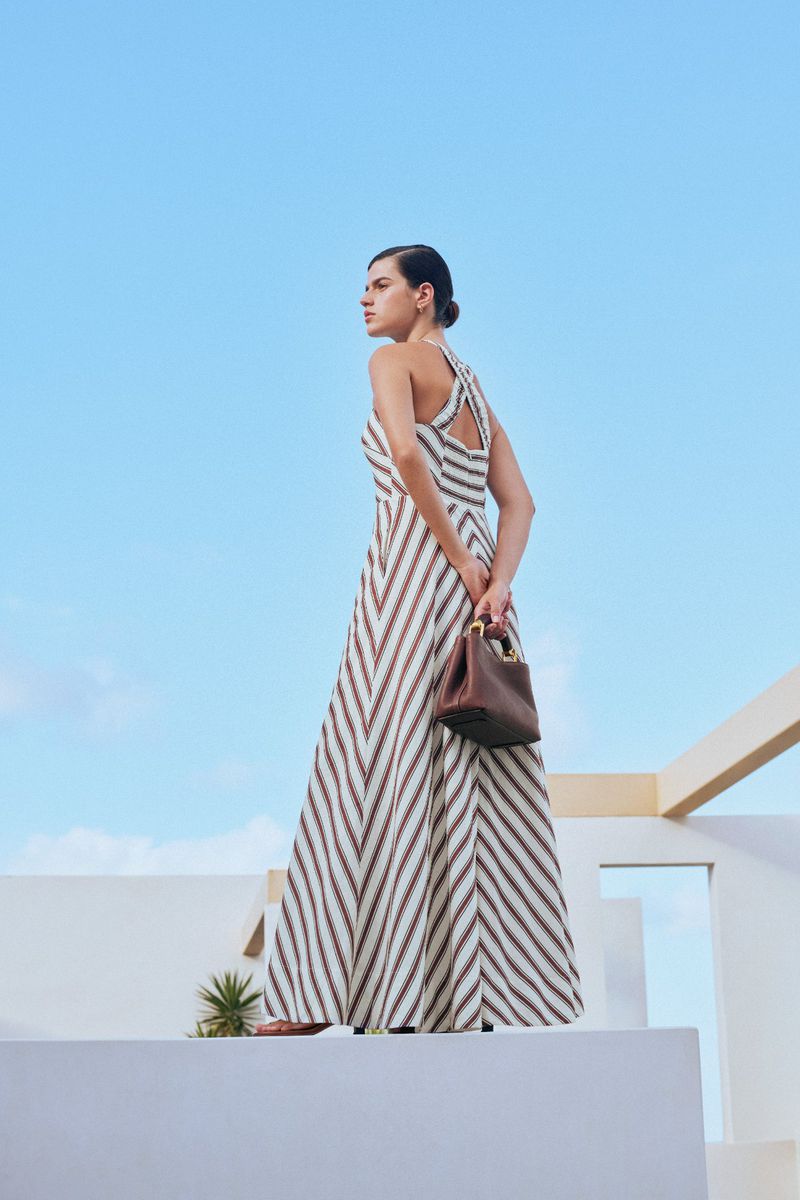 A woman in a striped maxi dress poses on a rooftop with a brown handbag, against a blue sky. Fashion, outdoor setting, clear weather.
