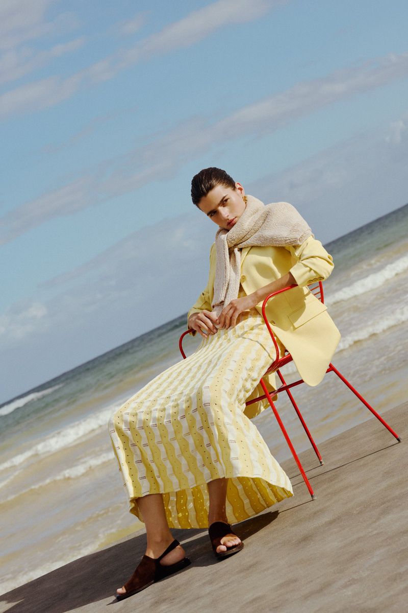 Woman poses on a red chair at the beach wearing a yellow blazer, patterned skirt, and scarf, ocean and blue sky background.