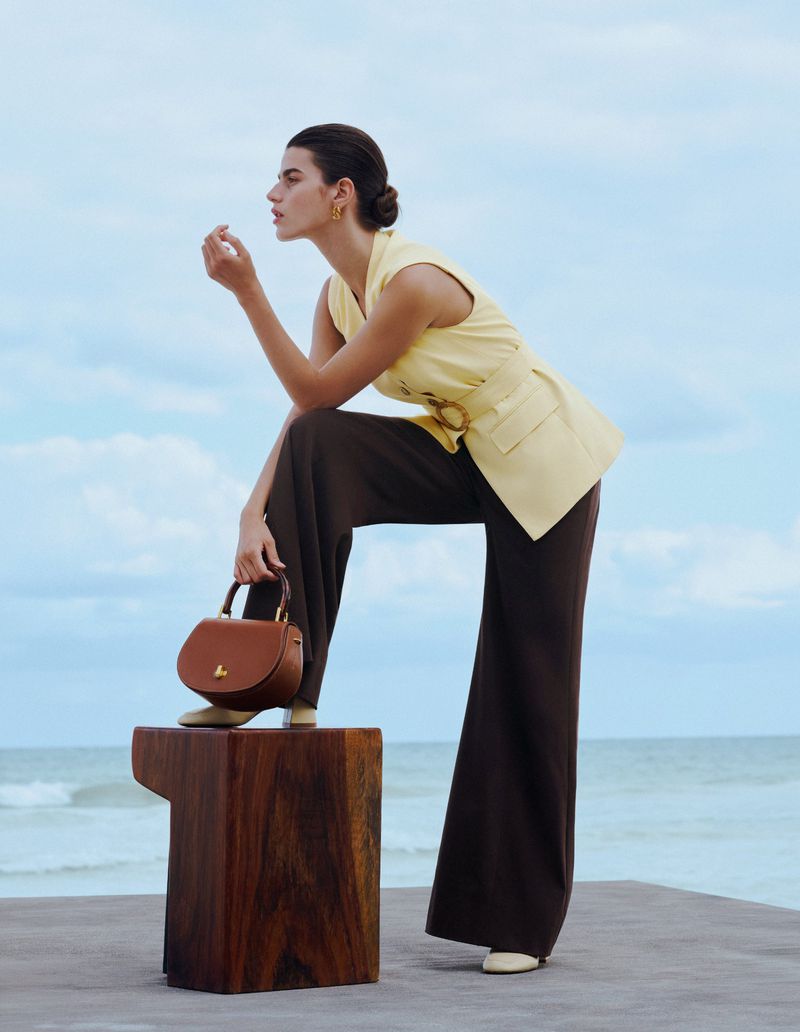 Adult woman in a yellow vest, wide-leg pants, and tan shoes posing with a brown handbag against a sky and ocean background.