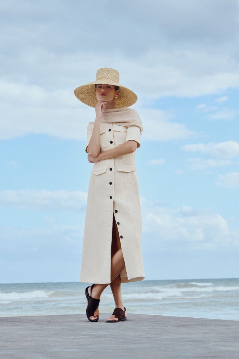Woman in a cream-colored dress and straw hat poses on a concrete pier with a blue sky and ocean in the background.