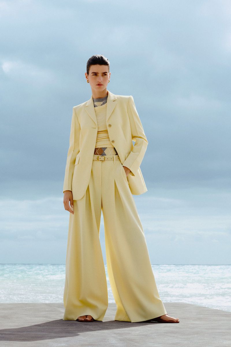 Woman in a pale yellow pantsuit posing in front of the ocean under a cloudy sky. Fashion shot featuring wide-leg trousers and blazer.