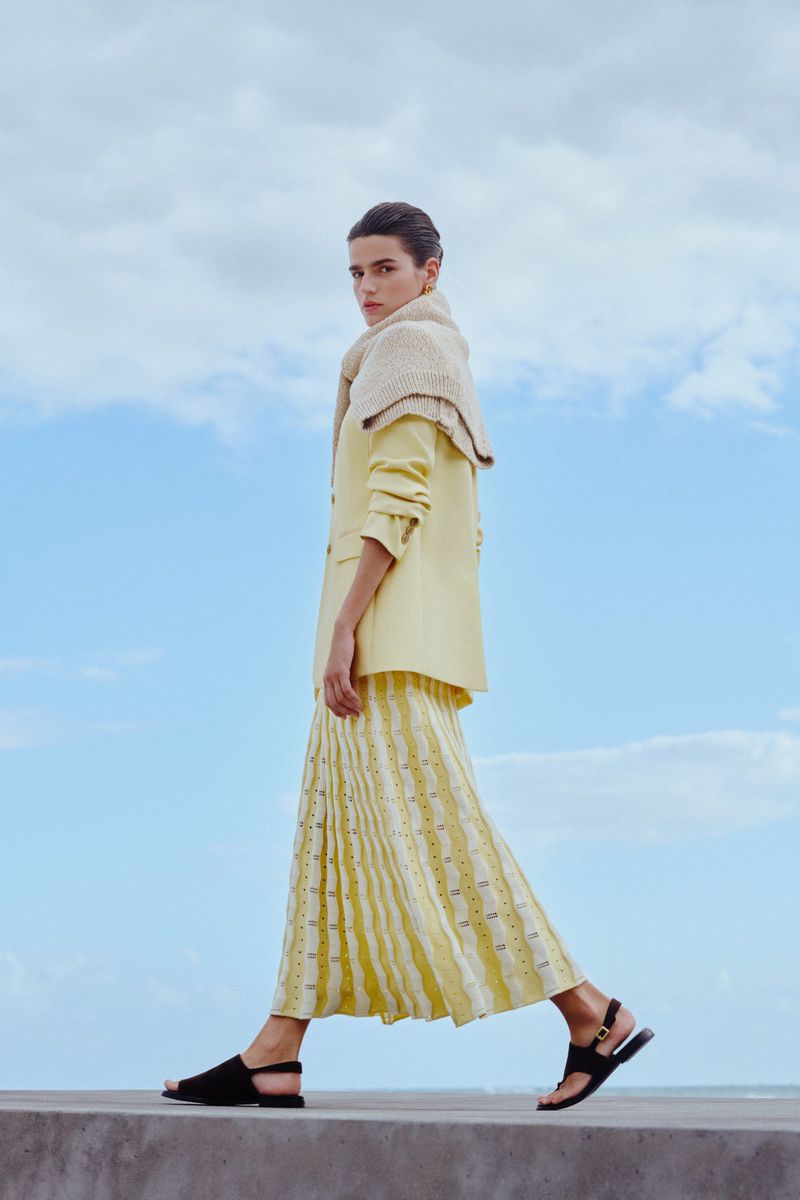 A woman walks across a concrete ledge in a yellow blazer, pleated yellow patterned midi skirt, and black sandals against a blue sky.