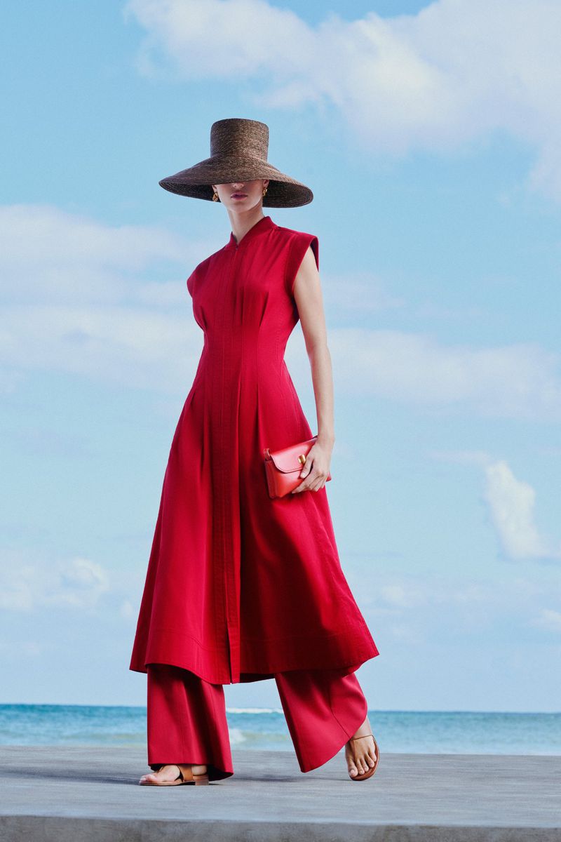 A woman wearing a red dress and hat stands on a concrete surface with a blue sky and ocean in the background for a fashion shot.