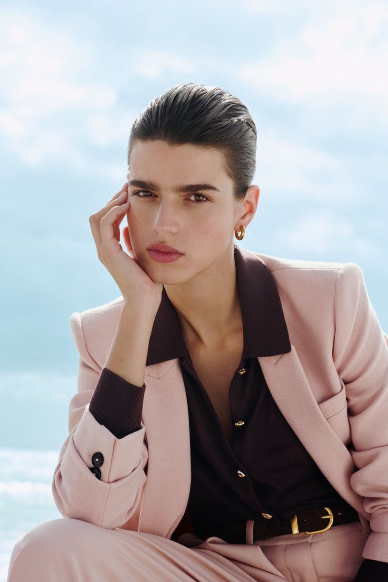 Portrait of an adult woman in a pink suit, styled with dark shirt and belt, posing against a bright blue sky with soft cloud cover.