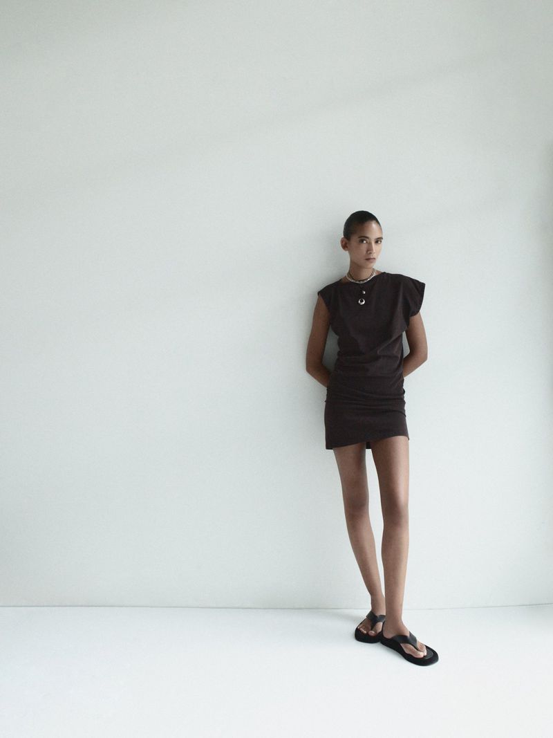 Model Summer Thompson standing against a white seamless backdrop wearing a dark sleeveless mini dress and black thong sandals for AGOLDE.