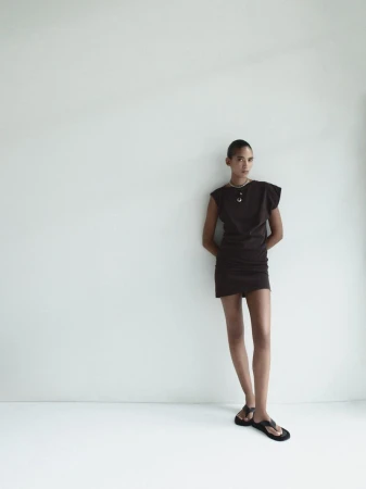 Model Summer Thompson standing against a white seamless backdrop wearing a dark sleeveless mini dress and black thong sandals for AGOLDE.