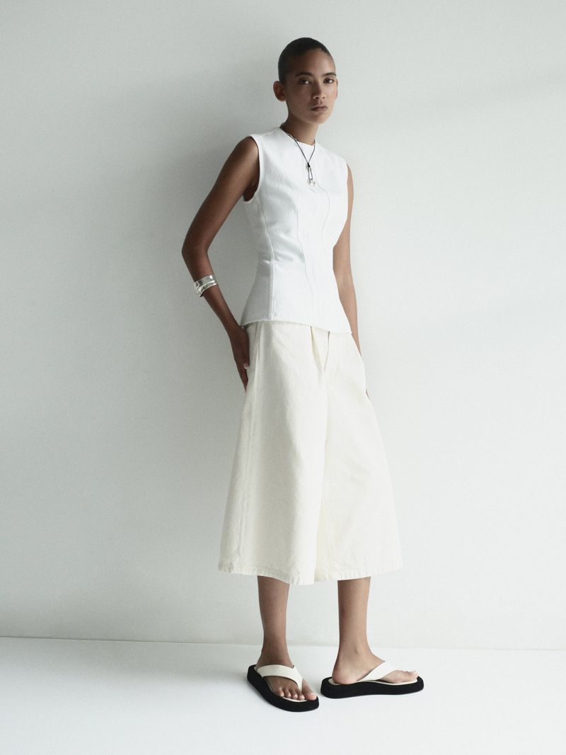 Full-length shot of an adult woman posing in a sleeveless white top, white culottes, and black sandals, against a neutral backdrop.