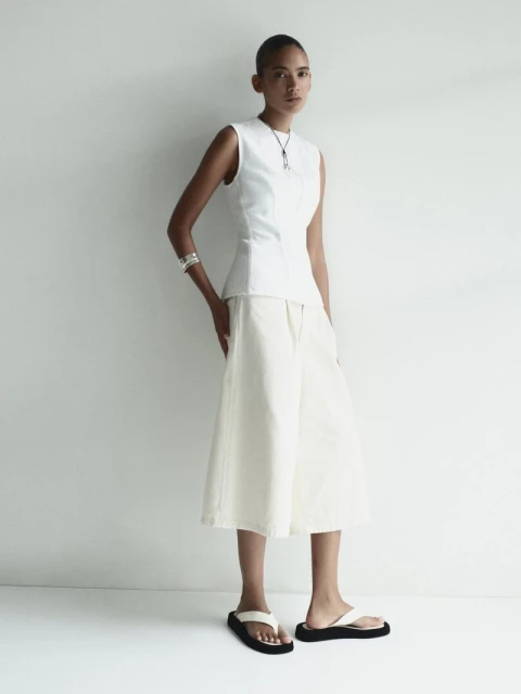 Full-length shot of an adult woman posing in a sleeveless white top, white culottes, and black sandals, against a neutral backdrop.