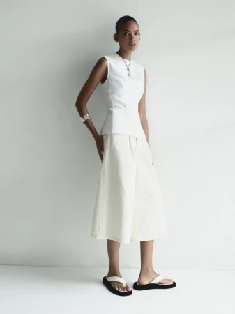 Full-length shot of an adult woman posing in a sleeveless white top, white culottes, and black sandals, against a neutral backdrop.