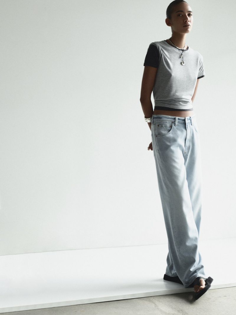 A slender woman with a shaved head models wide-leg light wash jeans and a gray t-shirt against a stark white backdrop in a fashion editorial.