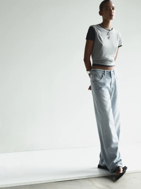 A slender woman with a shaved head models wide-leg light wash jeans and a gray t-shirt against a stark white backdrop in a fashion editorial.