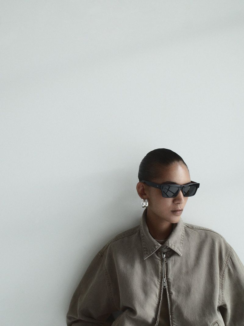 A woman with short hair and sunglasses poses in front of a white wall, wearing a beige jacket and silver hoop earrings, medium studio shot.