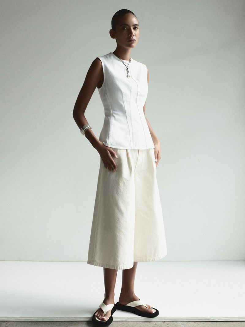 Full-shot of a light-skinned adult woman modeling a white sleeveless top with wide off-white culottes, standing in a neutral-toned studio.