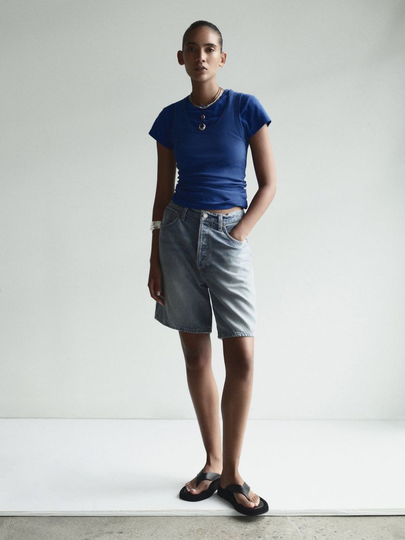 Model standing in a studio wearing a bright blue short-sleeved t-shirt, light wash denim shorts, and black thong sandals.
