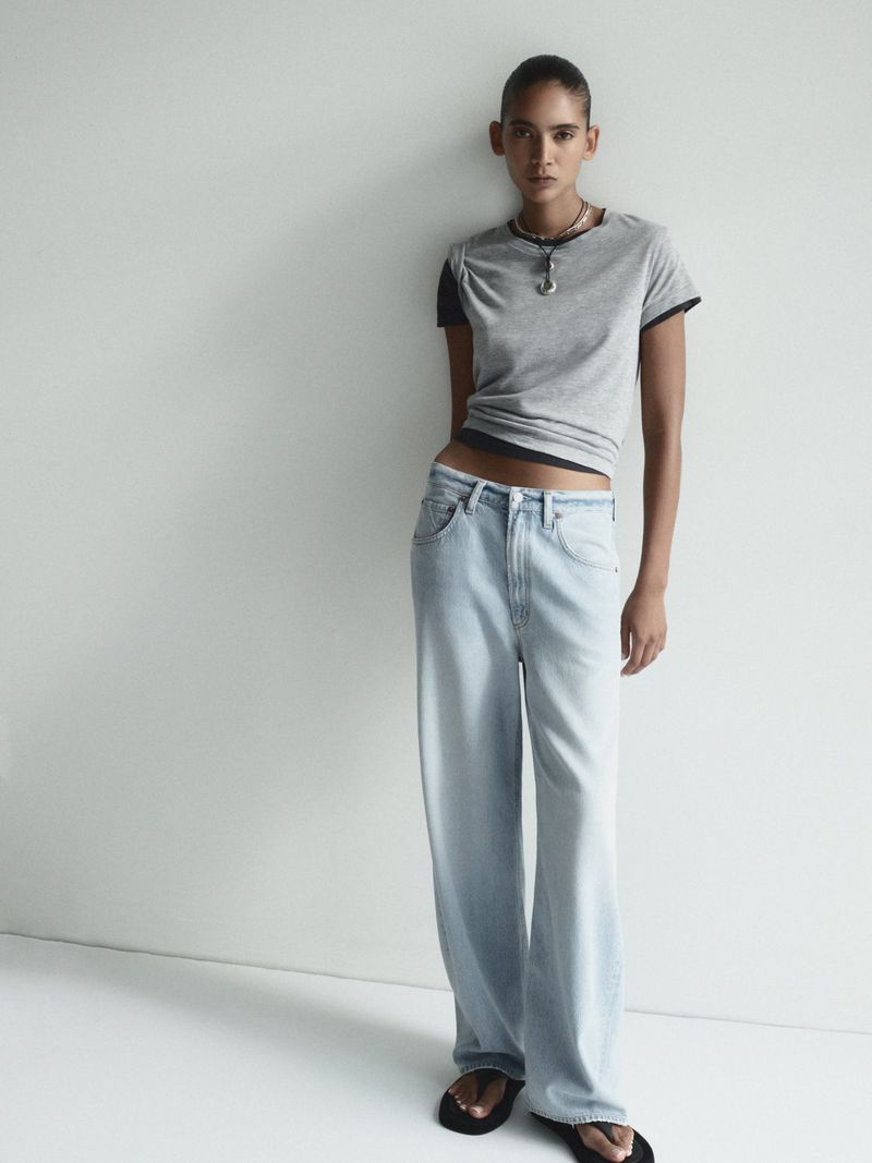 Model with pulled-back hair wearing a layered grey t-shirt and light-wash baggy jeans standing against a plain white wall