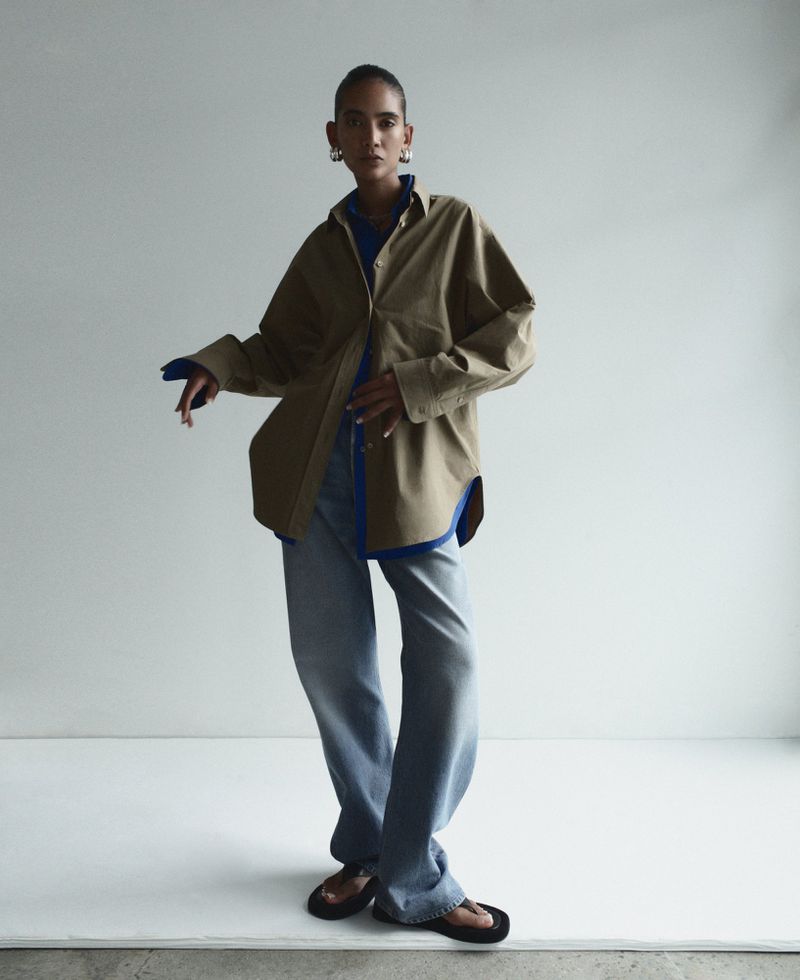 A woman in an olive-green shirt and jeans poses against a white background, her hands casually placed. Fashion editorial.