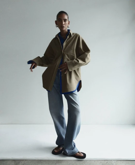 A woman in an olive-green shirt and jeans poses against a white background, her hands casually placed. Fashion editorial.