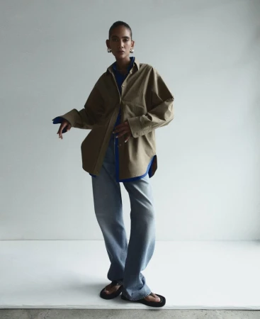 A woman in an olive-green shirt and jeans poses against a white background, her hands casually placed. Fashion editorial.
