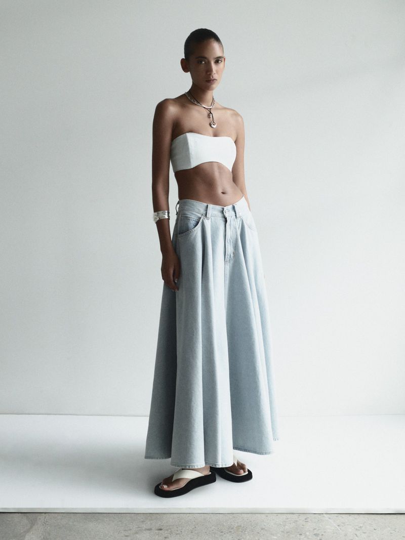 Woman in white tube top and light blue skirt poses in front of a white background in a fashion editorial with studio lighting.