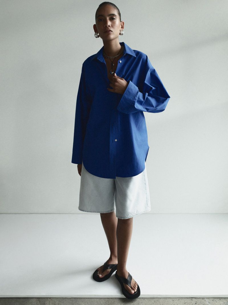 Model Summer Thompson wearing an oversized royal blue button-up shirt, light denim shorts, and black sandals in a white studio setting.
