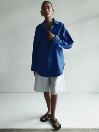 Model Summer Thompson wearing an oversized royal blue button-up shirt, light denim shorts, and black sandals in a white studio setting.