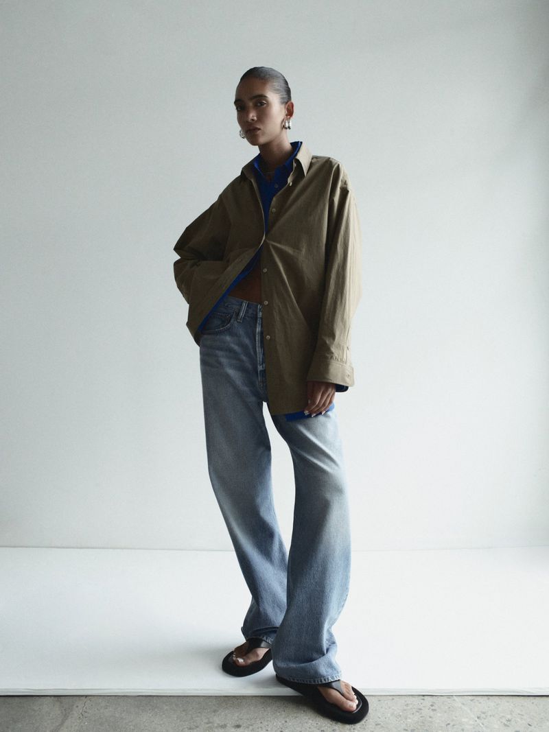 Woman poses in a studio wearing a tan button-down shirt, blue jeans, and black sandals, against a white backdrop with soft, diffused lighting.