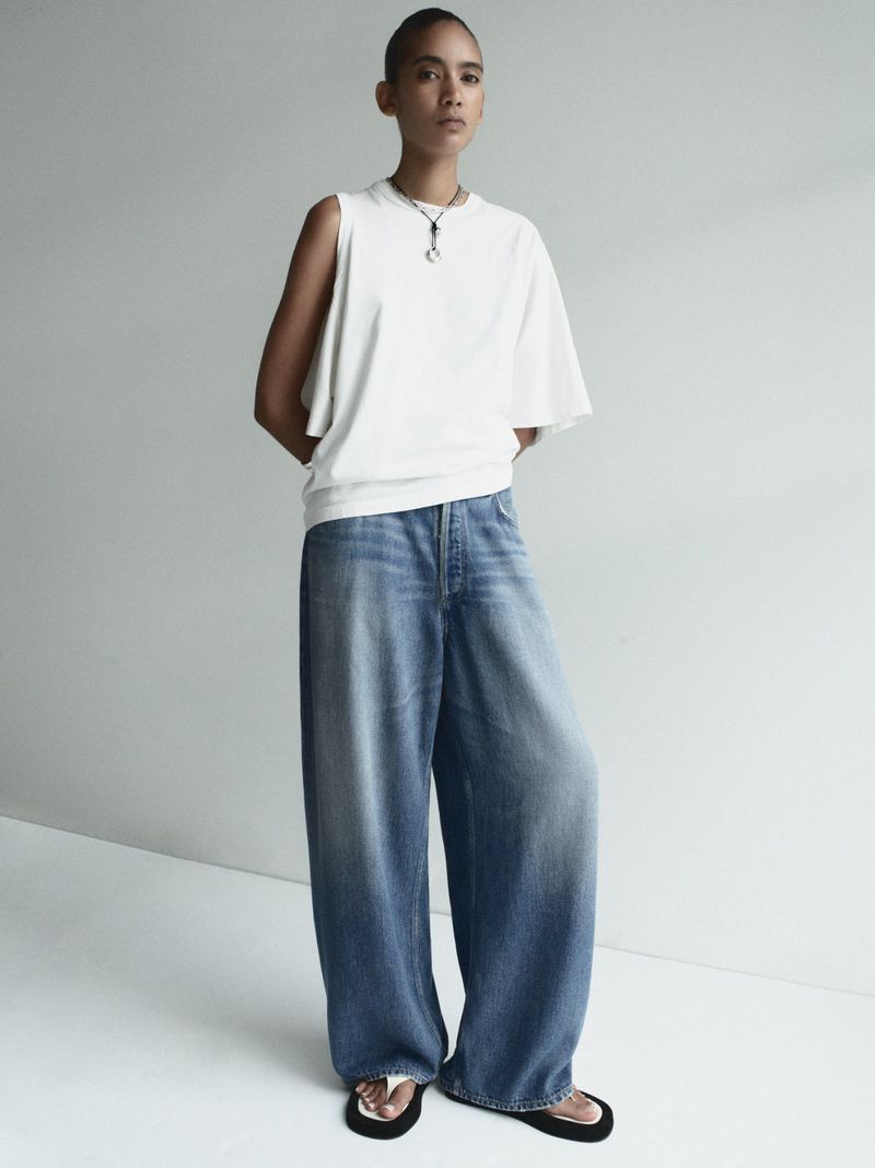 An adult woman poses indoors wearing a white, asymmetrical top, blue denim pants, and black slide sandals against a neutral background.